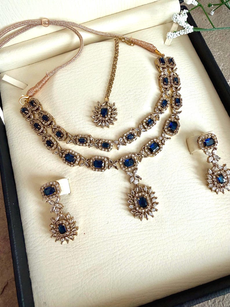 May include: A gold-toned jewelry set featuring a double-strand necklace with rectangular blue stones and clear crystals, a matching maang tikka with a central blue stone, and two drop earrings, each with a rectangular blue stone and crystal accents.