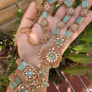 May include: A gold-toned necklace and matching earring set featuring rectangular and circular mint green stones, clear crystals, and small white pearl accents. The necklace includes a chain with a lobster clasp and a large circular pendant.
