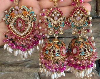 Może przedstawiać: Zestaw złotych kolczyków z dużym, ozdobnym sztyftem w kształcie serca oraz dwoma kolczykami typu jhumka w kształcie dzwonków. Biżuteria jest ozdobiona różowymi, pomarańczowymi, turkusowymi i zielonymi kamieniami oraz małymi białymi perłami.