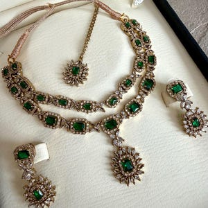 May include: A gold-toned jewelry set featuring a two-layered necklace with emerald green rectangular stones and clear crystals, a matching maang tikka with a single green stone, and two drop earrings with green stones and crystal accents.