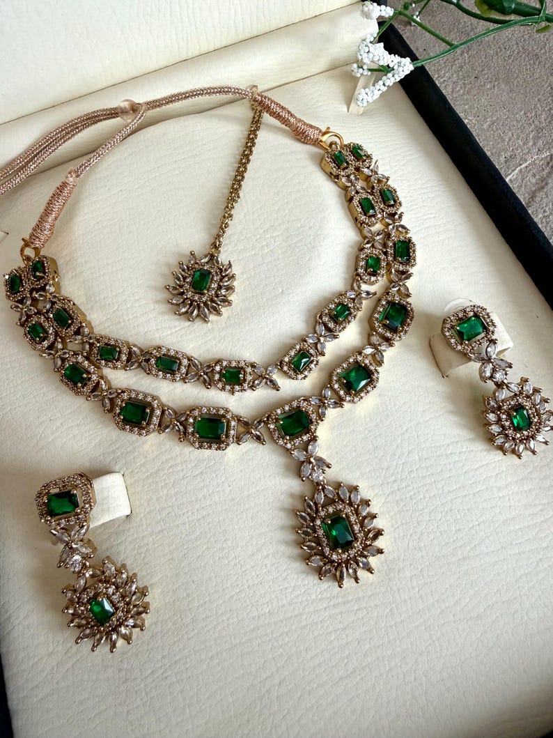 May include: A gold-toned jewelry set featuring a two-layered necklace with emerald green rectangular stones and clear crystals, a matching maang tikka with a single green stone, and two drop earrings with green stones and crystal accents.