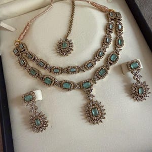 May include: A jewelry set featuring a double-strand necklace with rectangular light green stones and clear crystals, a matching maang tikka with a central light green stone, and two drop earrings with light green stones and clear crystal accents.
