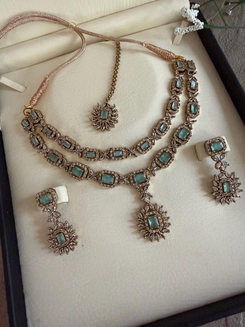May include: A jewelry set featuring a double-strand necklace with rectangular light green stones and clear crystals, a matching maang tikka with a central light green stone, and two drop earrings with light green stones and clear crystal accents.