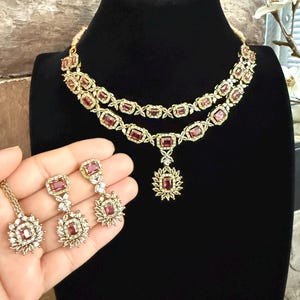 May include: A two-layer gold-tone necklace featuring rectangular pink stones surrounded by small clear crystals, with a matching pendant and a pair of drop earrings, each showcasing a rectangular pink stone and a floral-inspired crystal cluster.