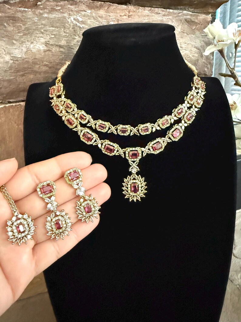 May include: A two-layer gold-tone necklace featuring rectangular pink stones surrounded by small clear crystals, with a matching pendant and a pair of drop earrings, each showcasing a rectangular pink stone and a floral-inspired crystal cluster.