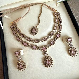 May include: A jewelry set featuring a double-layered necklace with rectangular light pink stones, a matching maang tikka, and two drop earrings. The pieces are crafted with gold-toned metal settings and small clear crystals surrounding each pink stone.