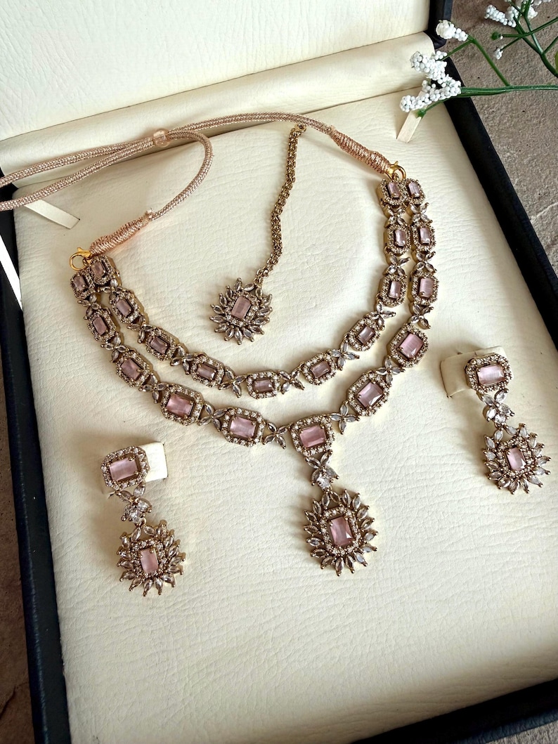 May include: A jewelry set featuring a double-layered necklace with rectangular light pink stones, a matching maang tikka, and two drop earrings. The pieces are crafted with gold-toned metal settings and small clear crystals surrounding each pink stone.