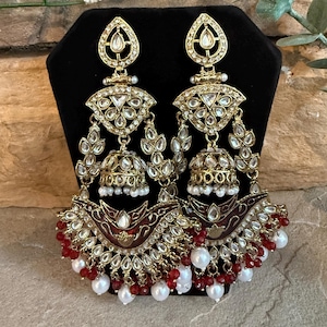 May include: A pair of gold-toned chandelier earrings featuring a teardrop-shaped top, a central bell-shaped jhumka element, and a large crescent-shaped base. The design is accented with clear stones, deep red enamel, and dangling red and white beads.