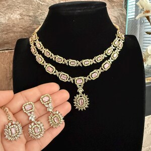 May include: A two-layer gold-tone necklace featuring rectangular light pink stones surrounded by small clear crystals. The set includes matching drop earrings, each with a rectangular light pink stone and a floral-shaped cluster of clear crystals.