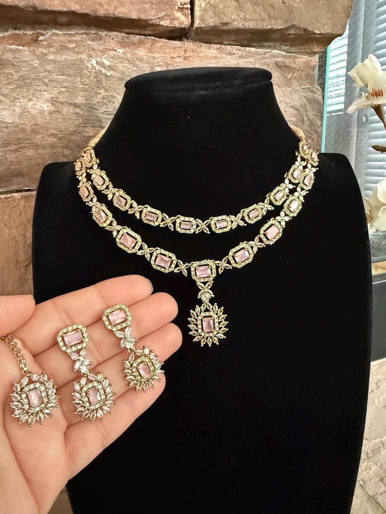 May include: A two-layer gold-tone necklace featuring rectangular light pink stones surrounded by small clear crystals. The set includes matching drop earrings, each with a rectangular light pink stone and a floral-shaped cluster of clear crystals.