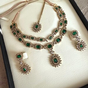 May include: A gold-toned jewelry set featuring a double-layered necklace with emerald-green rectangular stones and clear crystals, matching drop earrings with floral-shaped accents, and a coordinating maang tikka with a single green stone.