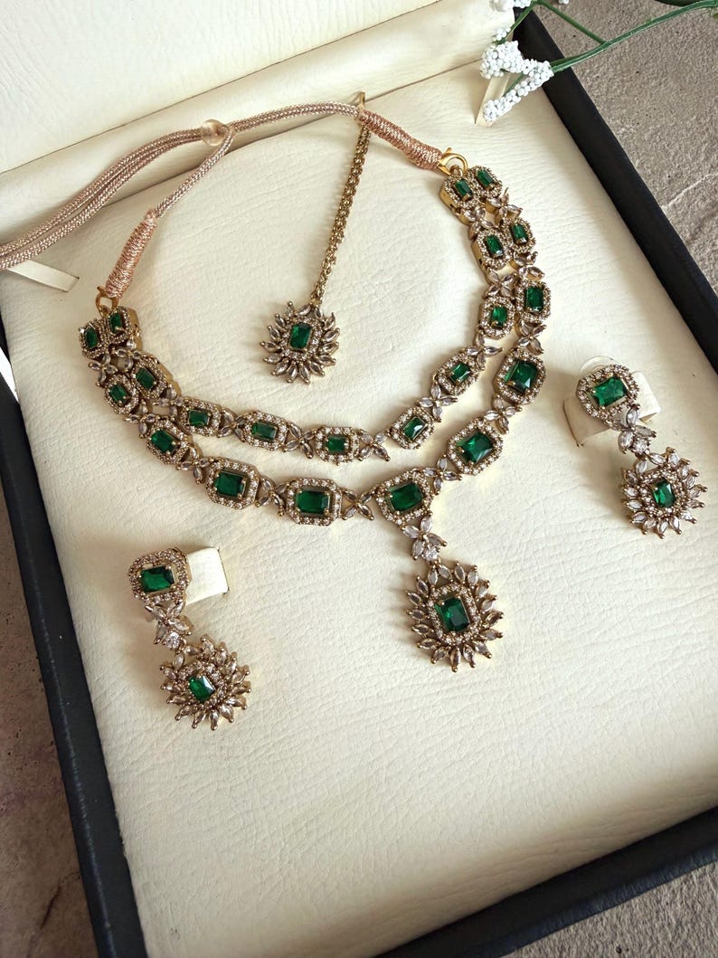 May include: A gold-toned jewelry set featuring a double-layered necklace with emerald-green rectangular stones and clear crystals, matching drop earrings with floral-shaped accents, and a coordinating maang tikka with a single green stone.