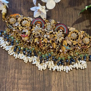 May include: Ornate gold-toned necklace with intricate floral and geometric designs. The necklace features colorful accents, including red, blue, and green, and is adorned with white pearl-like beads and dark blue and green crystal beads.