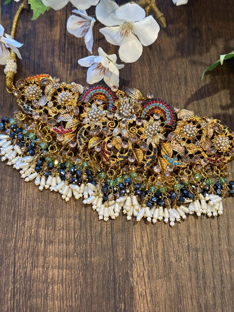 May include: Ornate gold-toned necklace with intricate floral and geometric designs. The necklace features colorful accents, including red, blue, and green, and is adorned with white pearl-like beads and dark blue and green crystal beads.