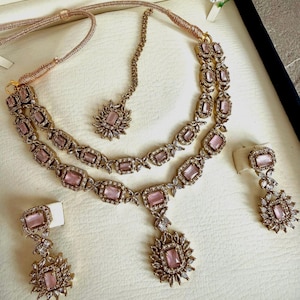May include: A gold-toned jewelry set featuring a double-strand necklace with rectangular pink stones and clear crystal accents, a matching maang tikka with a central pink stone, and two drop earrings with rectangular pink stones and crystal clusters.