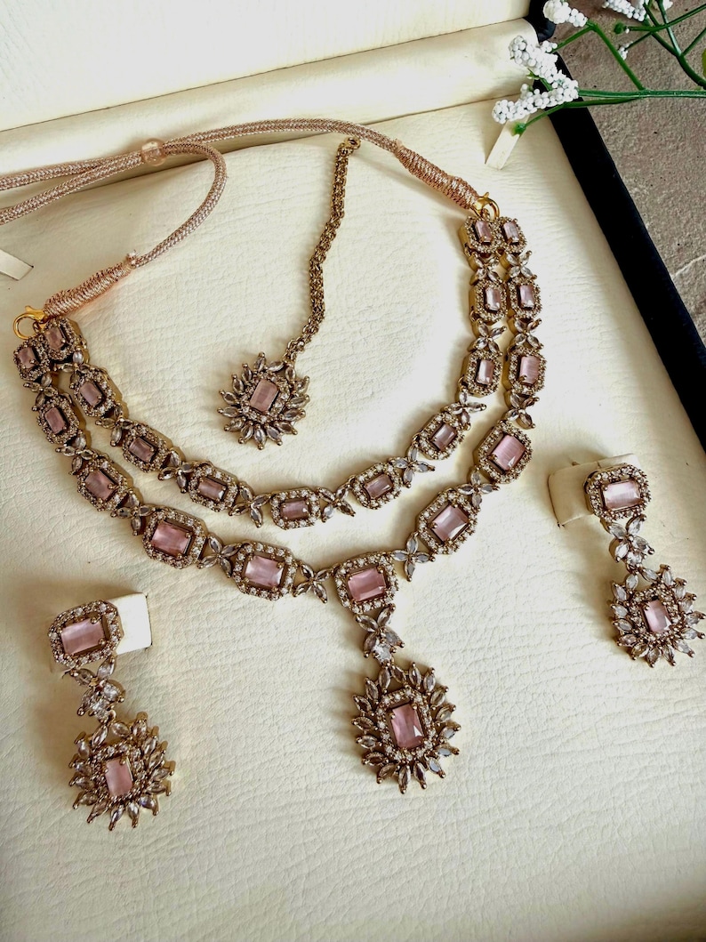 May include: A gold-toned jewelry set featuring a double-strand necklace with rectangular pink stones and clear crystal accents, a matching maang tikka with a central pink stone, and two drop earrings with rectangular pink stones and crystal clusters.