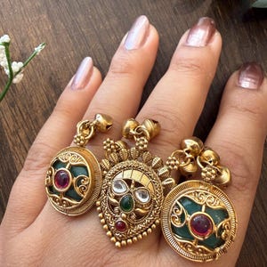 May include: Three ornate gold-toned rings with intricate detailing. Each ring features a central gemstone, with a deep green or red hue, set within a decorative frame. The rings are displayed on a hand.