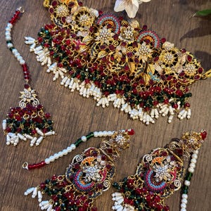 May include: An ornate gold-toned jewelry set featuring a necklace, earrings, and a headpiece. The set is adorned with red and green beads, white pearls, and intricate floral designs. The jewelry is displayed on a wooden surface.