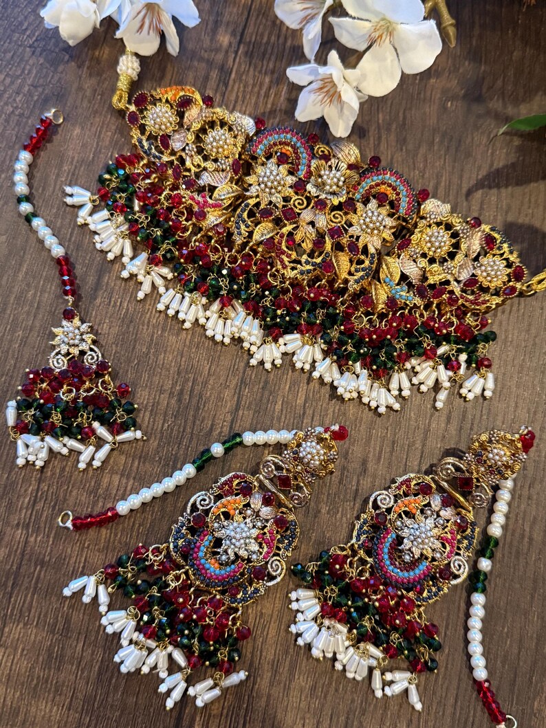 May include: An ornate gold-toned jewelry set featuring a necklace, earrings, and a headpiece. The set is adorned with red and green beads, white pearls, and intricate floral designs. The jewelry is displayed on a wooden surface.