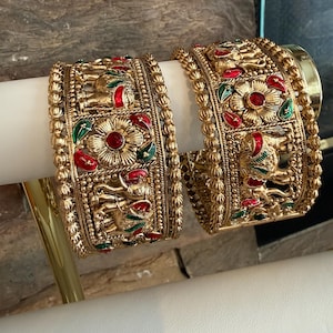 May include: Two gold-colored bangles with intricate designs. Each bangle features floral and elephant motifs, accented with red and green enamel. The bangles are displayed on a white surface, showcasing their ornate details and craftsmanship.