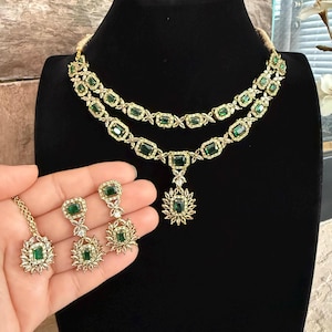 May include: A gold-toned jewelry set featuring a two-layered necklace and matching earrings. The pieces are adorned with rectangular emerald green stones surrounded by small clear crystals. The necklace includes a central pendant with a floral design.