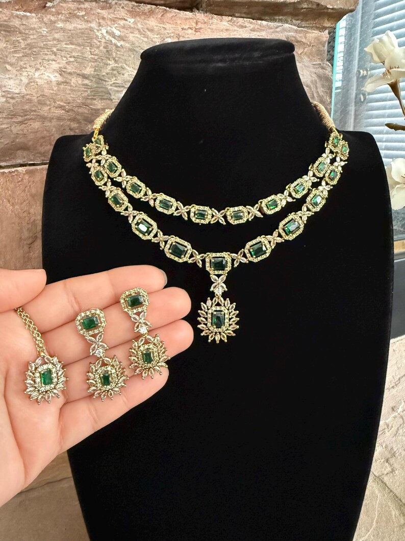 May include: A gold-toned jewelry set featuring a two-layered necklace and matching earrings. The pieces are adorned with rectangular emerald green stones surrounded by small clear crystals. The necklace includes a central pendant with a floral design.