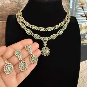 May include: A two-layered gold-tone necklace and matching drop earrings set featuring rectangular light green stones surrounded by small clear white crystals. The necklace includes a central pendant with a matching light green stone.