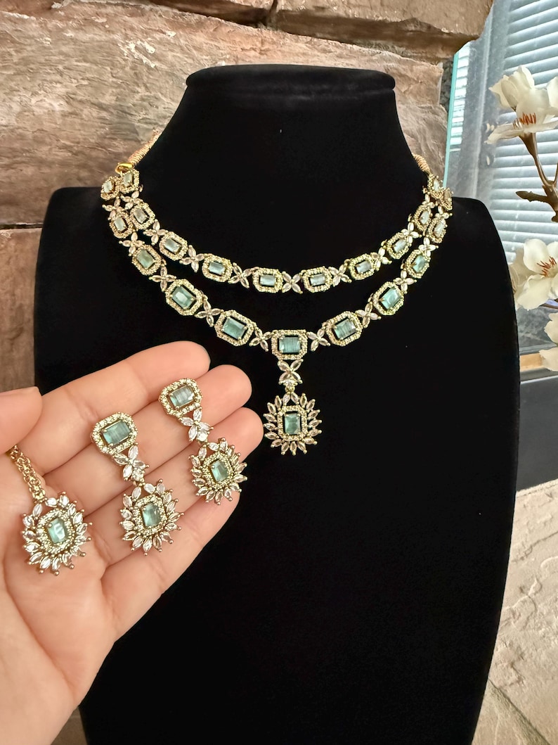 May include: A two-layered gold-tone necklace and matching drop earrings set featuring rectangular light green stones surrounded by small clear white crystals. The necklace includes a central pendant with a matching light green stone.