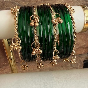 May include: A set of green glass bangles with gold-colored accents. The bangles are adorned with dangling gold-colored charms. The bangles are displayed on a white and gold surface, with a textured brown background.