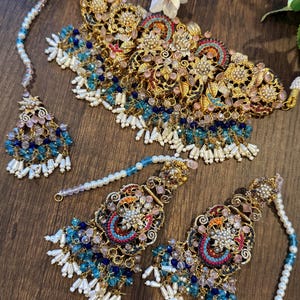 May include: An ornate gold-toned jewelry set featuring a necklace, earrings, and a headpiece. The set is adorned with blue, pink, and white beads, along with pearl accents. The intricate design includes floral and geometric patterns, creating a luxurious aesthetic.