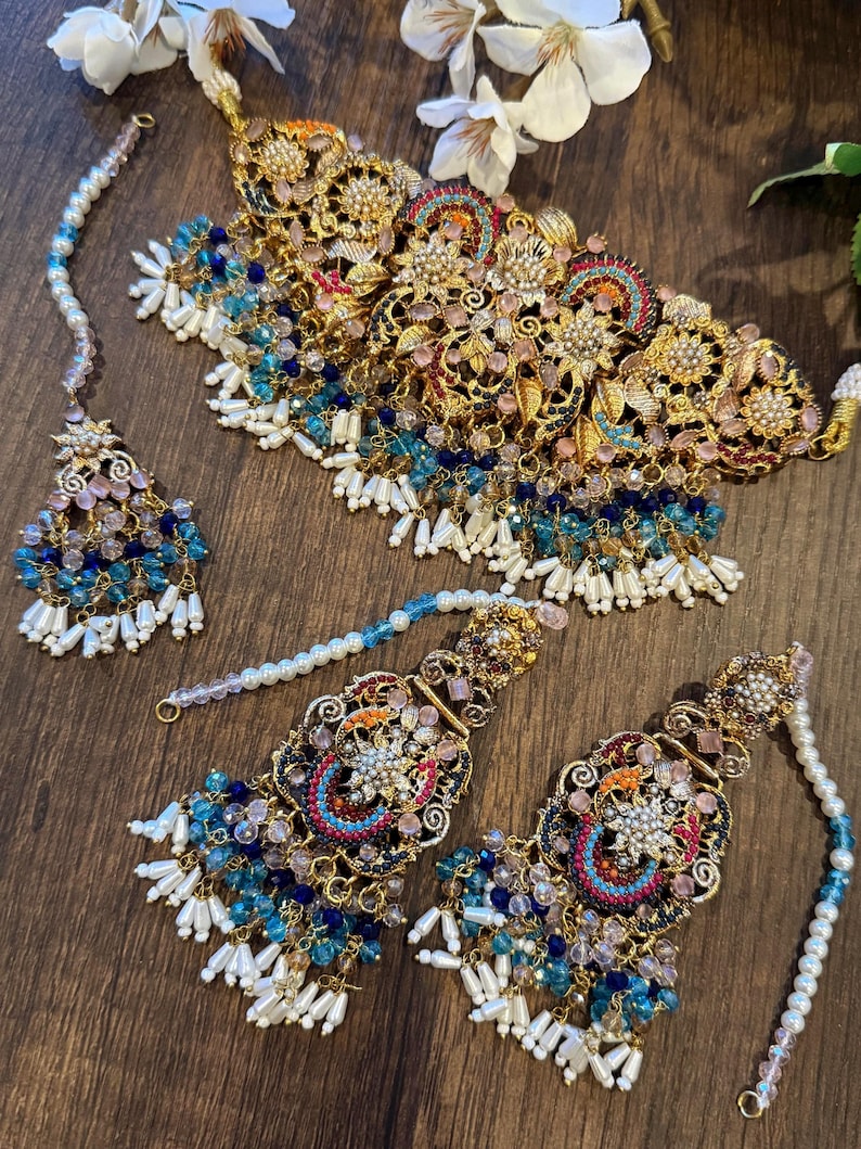 May include: An ornate gold-toned jewelry set featuring a necklace, earrings, and a headpiece. The set is adorned with blue, pink, and white beads, along with pearl accents. The intricate design includes floral and geometric patterns, creating a luxurious aesthetic.