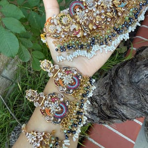May include: An ornate gold-toned jewelry set featuring a necklace, earrings, and a bracelet. The pieces are adorned with colorful gemstones, white pearls, and intricate floral designs. The set is displayed against a backdrop of green foliage.