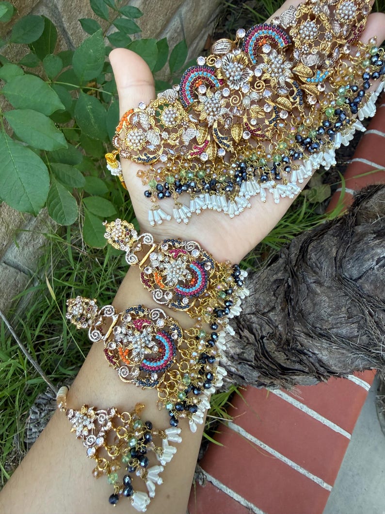 May include: An ornate gold-toned jewelry set featuring a necklace, earrings, and a bracelet. The pieces are adorned with colorful gemstones, white pearls, and intricate floral designs. The set is displayed against a backdrop of green foliage.