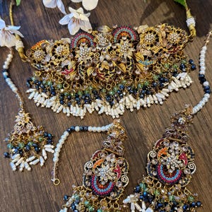 May include: An ornate gold-toned jewelry set featuring a necklace, earrings, and a headpiece. The set is embellished with colorful gemstones, pearls, and intricate filigree designs. The necklace and earrings have dangling beaded accents.