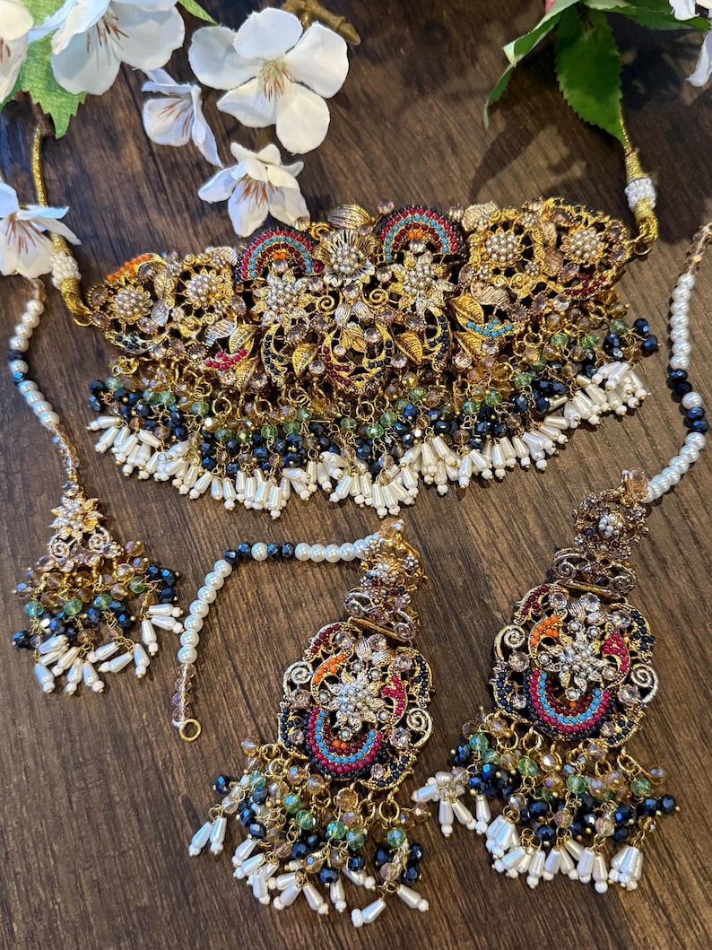 May include: An ornate gold-toned jewelry set featuring a necklace, earrings, and a headpiece. The set is embellished with colorful gemstones, pearls, and intricate filigree designs. The necklace and earrings have dangling beaded accents.