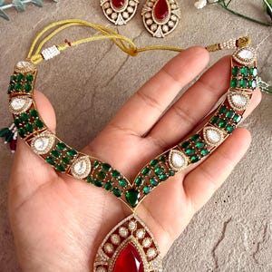 May include: A gold-toned necklace and matching earrings set. The necklace features emerald green and clear gemstones, with a large teardrop-shaped red gemstone pendant. The earrings have a similar design with red gemstones.