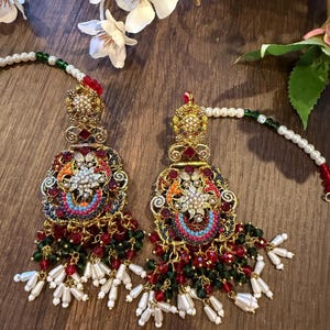 May include: A pair of ornate, gold-toned earrings with intricate detailing. The earrings feature a floral design with red, green, and blue accents, and are adorned with pearl and bead dangles. The earrings are set against a wooden background.