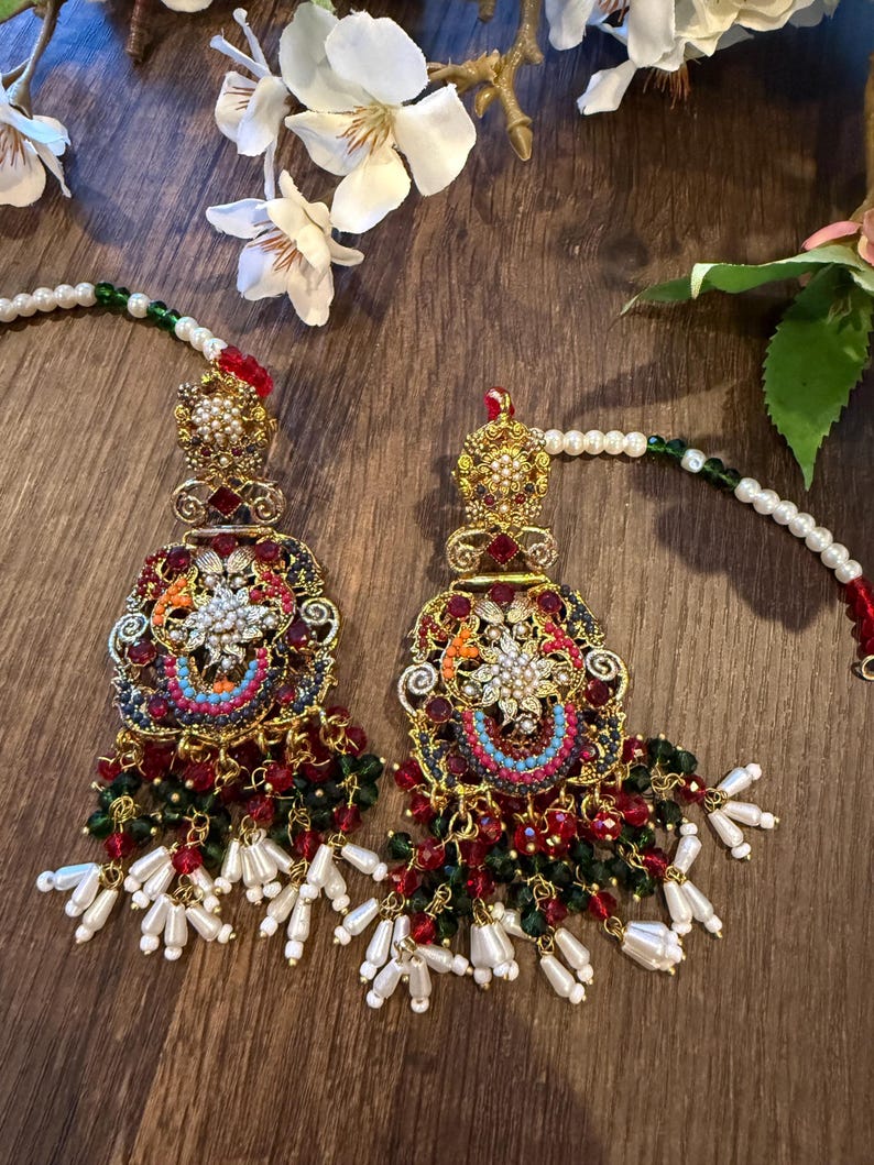 May include: A pair of ornate, gold-toned earrings with intricate detailing. The earrings feature a floral design with red, green, and blue accents, and are adorned with pearl and bead dangles. The earrings are set against a wooden background.