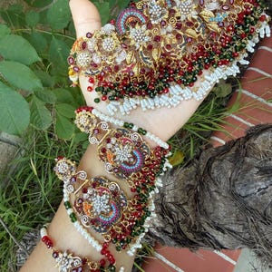 May include: Ornate gold-toned jewelry set featuring a necklace, earrings, and a bracelet. The pieces are adorned with red, green, and white beads, along with intricate floral and geometric designs. The jewelry is displayed against a natural green background.