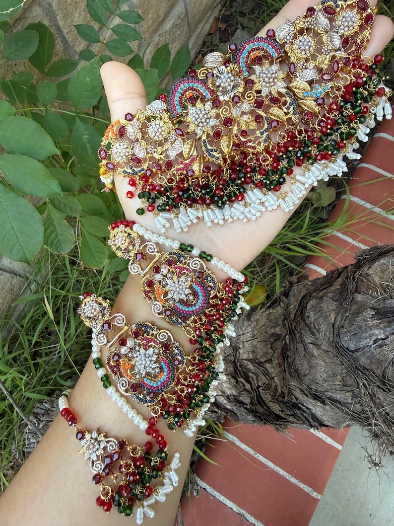 May include: Ornate gold-toned jewelry set featuring a necklace, earrings, and a bracelet. The pieces are adorned with red, green, and white beads, along with intricate floral and geometric designs. The jewelry is displayed against a natural green background.