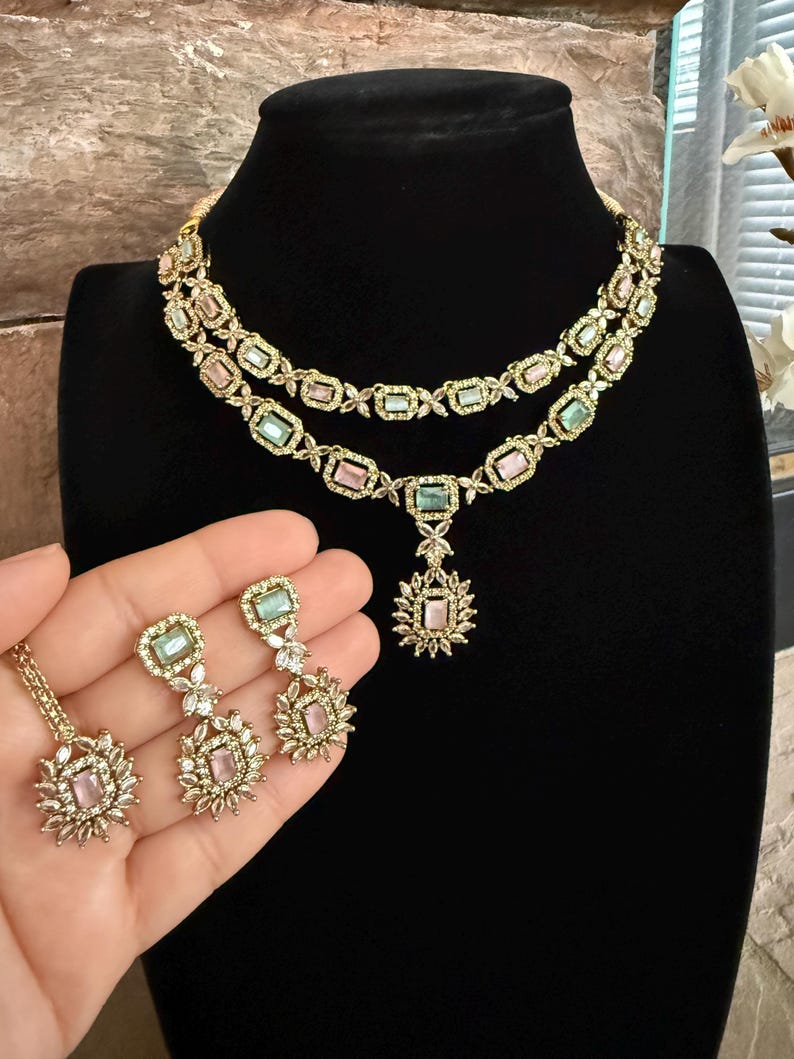 May include: A two-layer gold-tone necklace featuring alternating rectangular pastel pink and mint green stones set in ornate metal frames, with a matching pendant and a pair of drop earrings showcasing similar rectangular stones and floral-inspired accents.