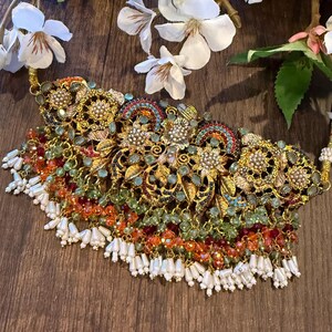 May include: An ornate gold-toned necklace with intricate floral and filigree designs. The necklace features colorful beads in shades of green, orange, and red, with white beaded fringe. White flowers are in the background.