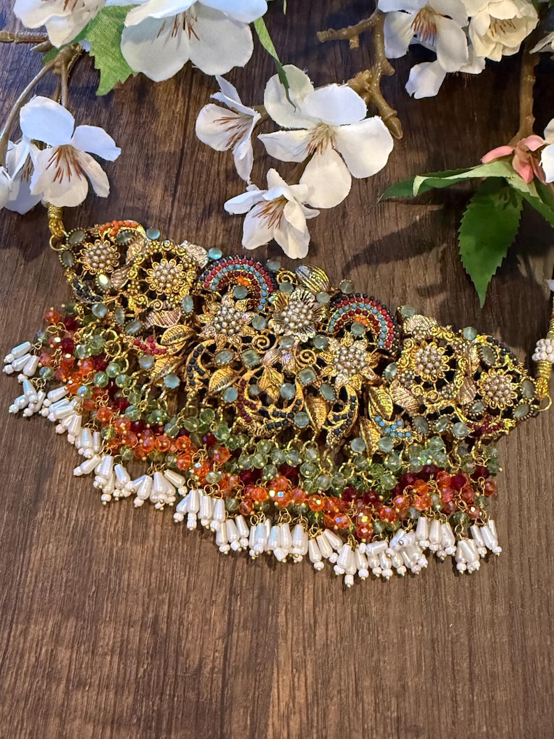 May include: An ornate gold-toned necklace with intricate floral and filigree designs. The necklace features colorful beads in shades of green, orange, and red, with white beaded fringe. White flowers are in the background.