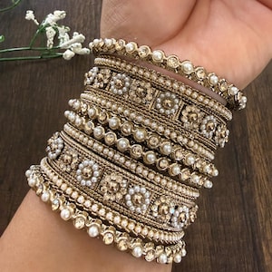 May include: A stack of gold-toned metal bangles featuring intricate floral patterns, embedded with small white faux pearls and clear sparkling crystals. The set consists of multiple bands with varying textures, including beaded edges and filigree details.