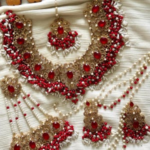 May include: A gold-toned bridal jewelry set featuring a necklace, earrings, and headpiece. The set is adorned with red gemstones and small red and white beads. The jewelry is displayed on a white fabric background.