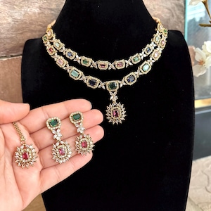 May include: A two-layered gold-tone necklace featuring rectangular multi-colored gemstones and clear accent stones, paired with matching drop earrings and a single pendant. The jewelry set displays emerald, sapphire, and ruby-colored stones.