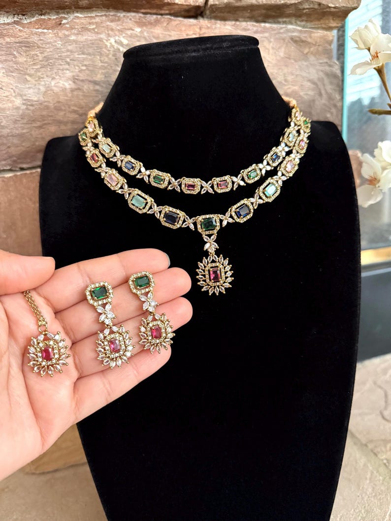 May include: A two-layered gold-tone necklace featuring rectangular multi-colored gemstones and clear accent stones, paired with matching drop earrings and a single pendant. The jewelry set displays emerald, sapphire, and ruby-colored stones.