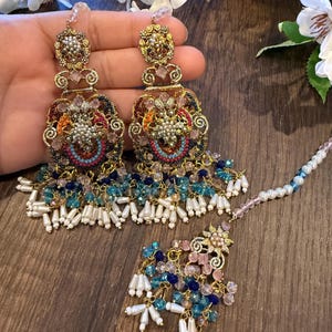 May include: Ornate gold-toned earrings and a matching headpiece. The earrings feature intricate filigree, colorful beadwork in shades of blue, pink, and orange, and dangling white pearl-like beads. The headpiece has a floral design and a beaded chain.