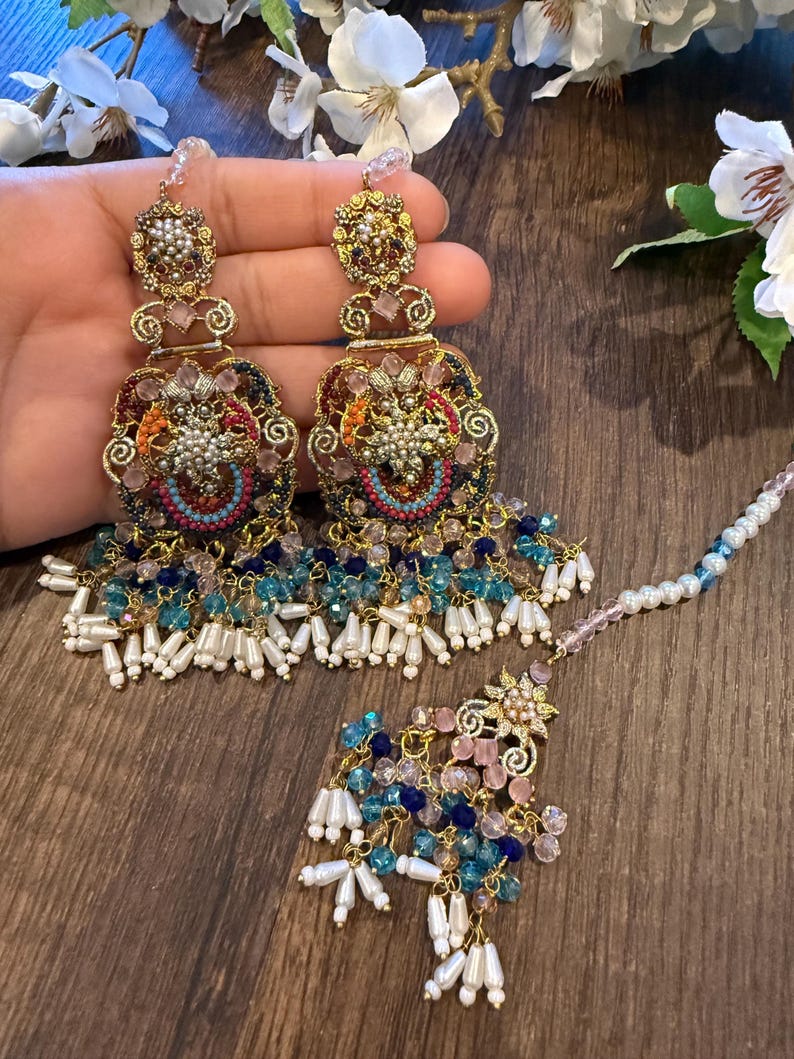 May include: Ornate gold-toned earrings and a matching headpiece. The earrings feature intricate filigree, colorful beadwork in shades of blue, pink, and orange, and dangling white pearl-like beads. The headpiece has a floral design and a beaded chain.