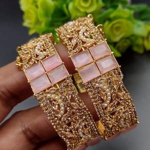 May include: Two gold bangles with pink glass stones and clear rhinestones. The bangles are decorated with a floral design.